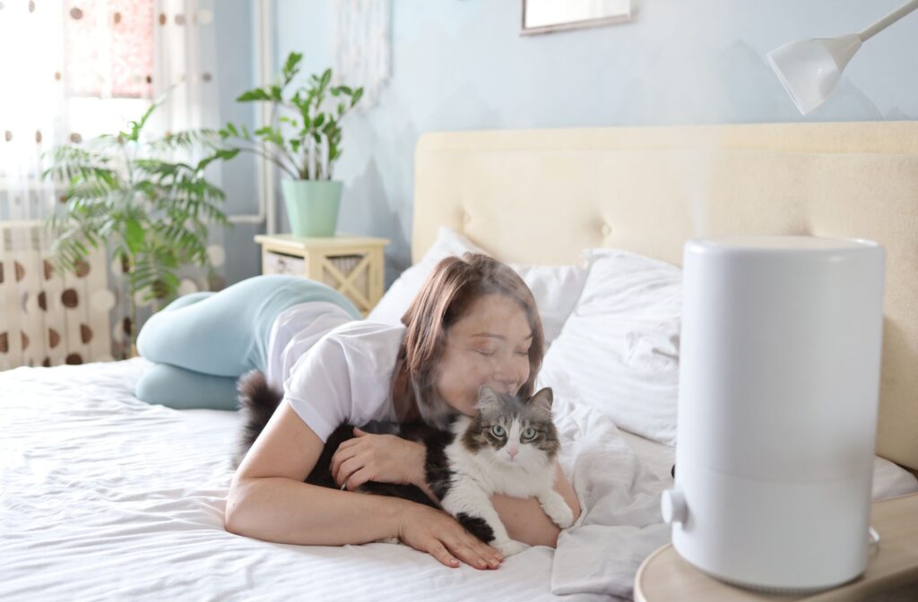 A person snuggles with their cat on the bed while enjoying the benefits of a humidifier for dry eyes.